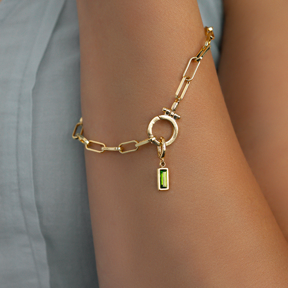 Gold chain bracelet with a green gemstone charm on a wrist against a neutral background