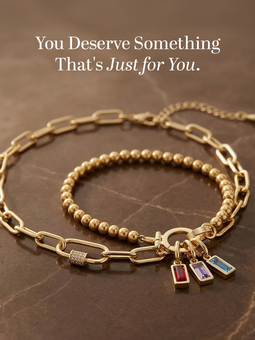 Gold bracelet with charms on a brown surface with text 'You Deserve Something That's Just for You'.