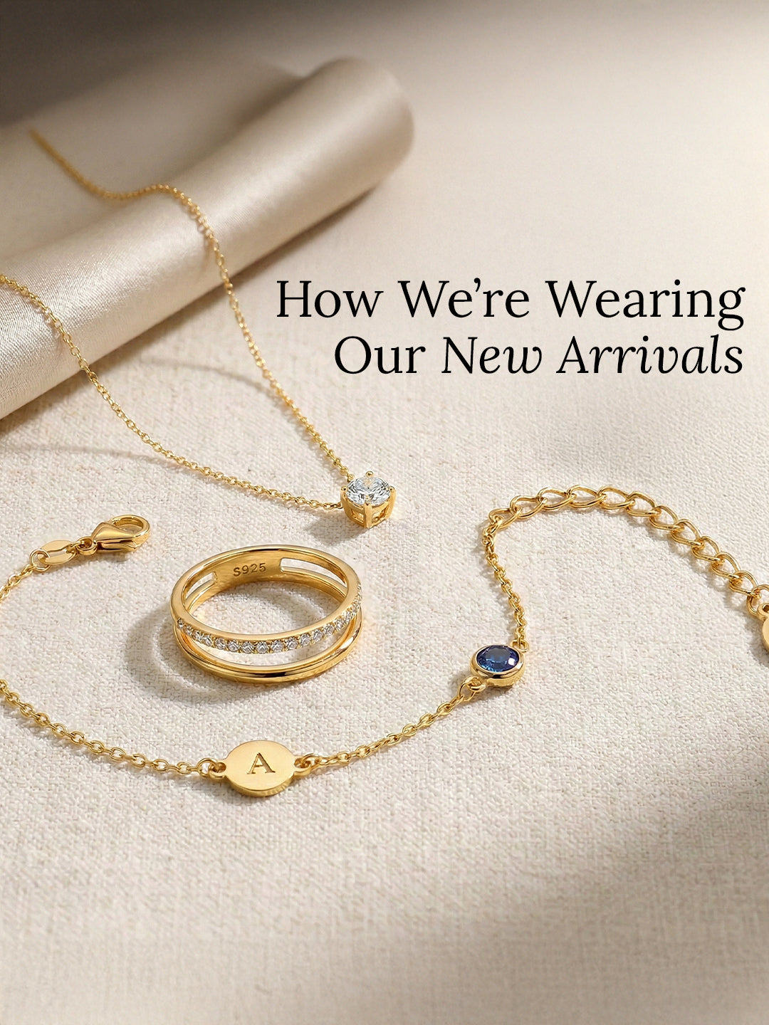 Gold jewelry including a bracelet, ring, and necklace on a beige surface with text How We're Wearing Our New Arrivals.