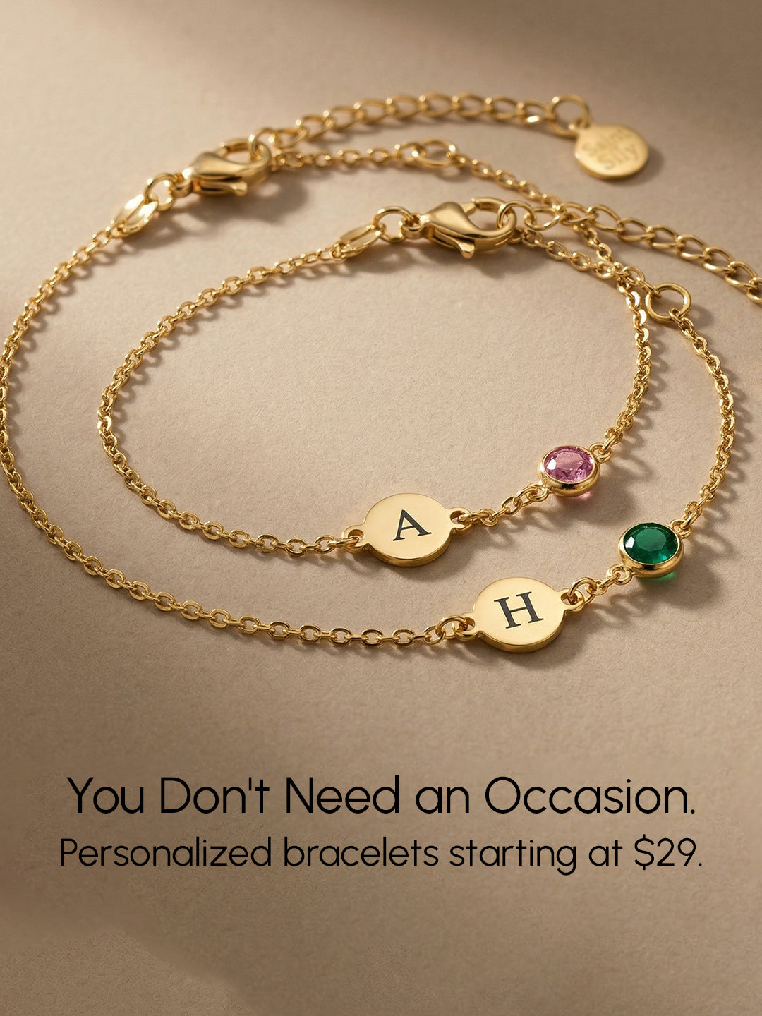 Two personalized gold bracelets with initial charms and pink and green gemstones on a beige background above the text: “You Don't Need an Occasion. Personalized bracelets starting at $29.
