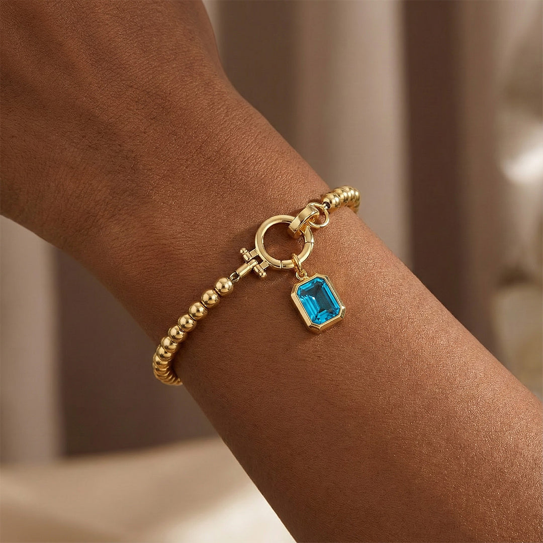 Gold bracelet with a blue gemstone charm on a wrist against a neutral background