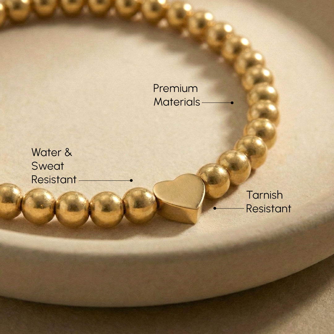 Gold beaded bracelet with a heart charm on a beige surface, labeled for features.