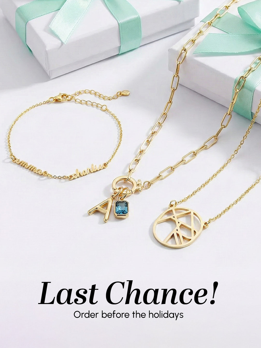 Gold necklace with peace sign charm and blue gemstone, gold bracelet, and white gift box with green ribbon on a light gray background.
