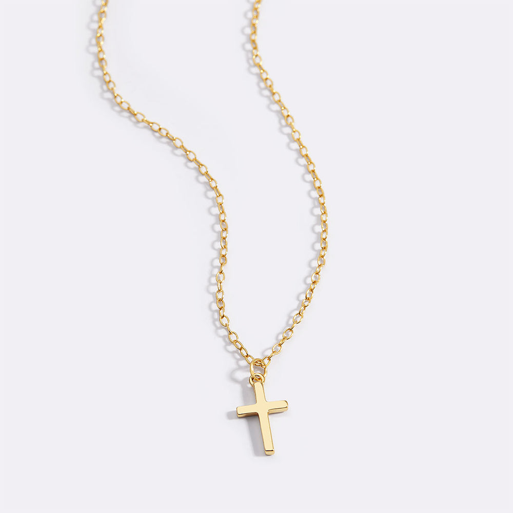 A gold-colored cross necklace with a dainty cable chain laid out against a plain background.