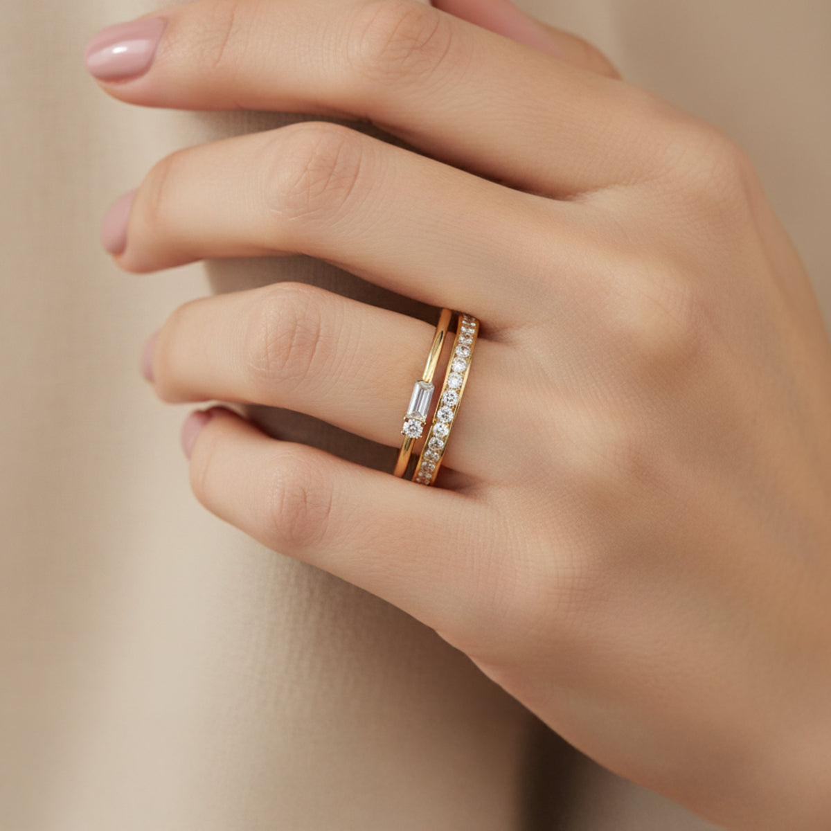Hand wearing two gold rings with diamond accents on a neutral background