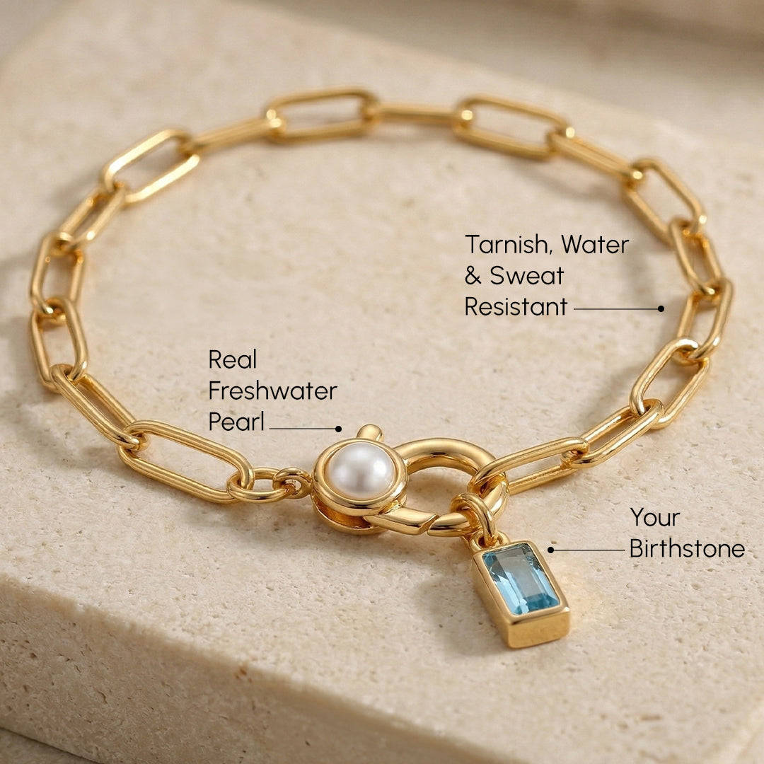 Gold chain bracelet with a pearl and gemstone charm on a beige stone surface
