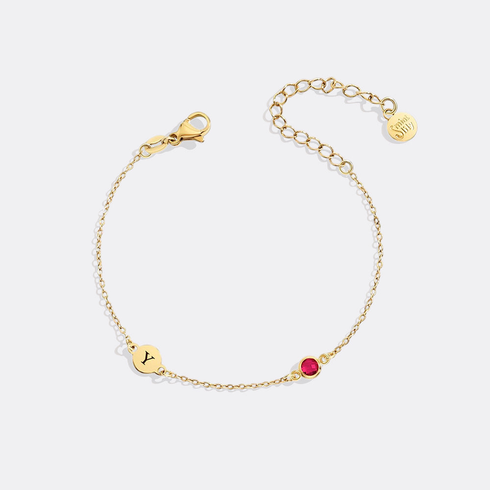 Gold bracelet with a red gemstone and gold tag on a white background