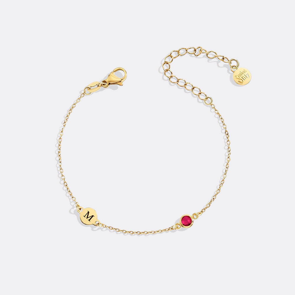 Gold bracelet with a red gemstone and 'M' charm on a white background