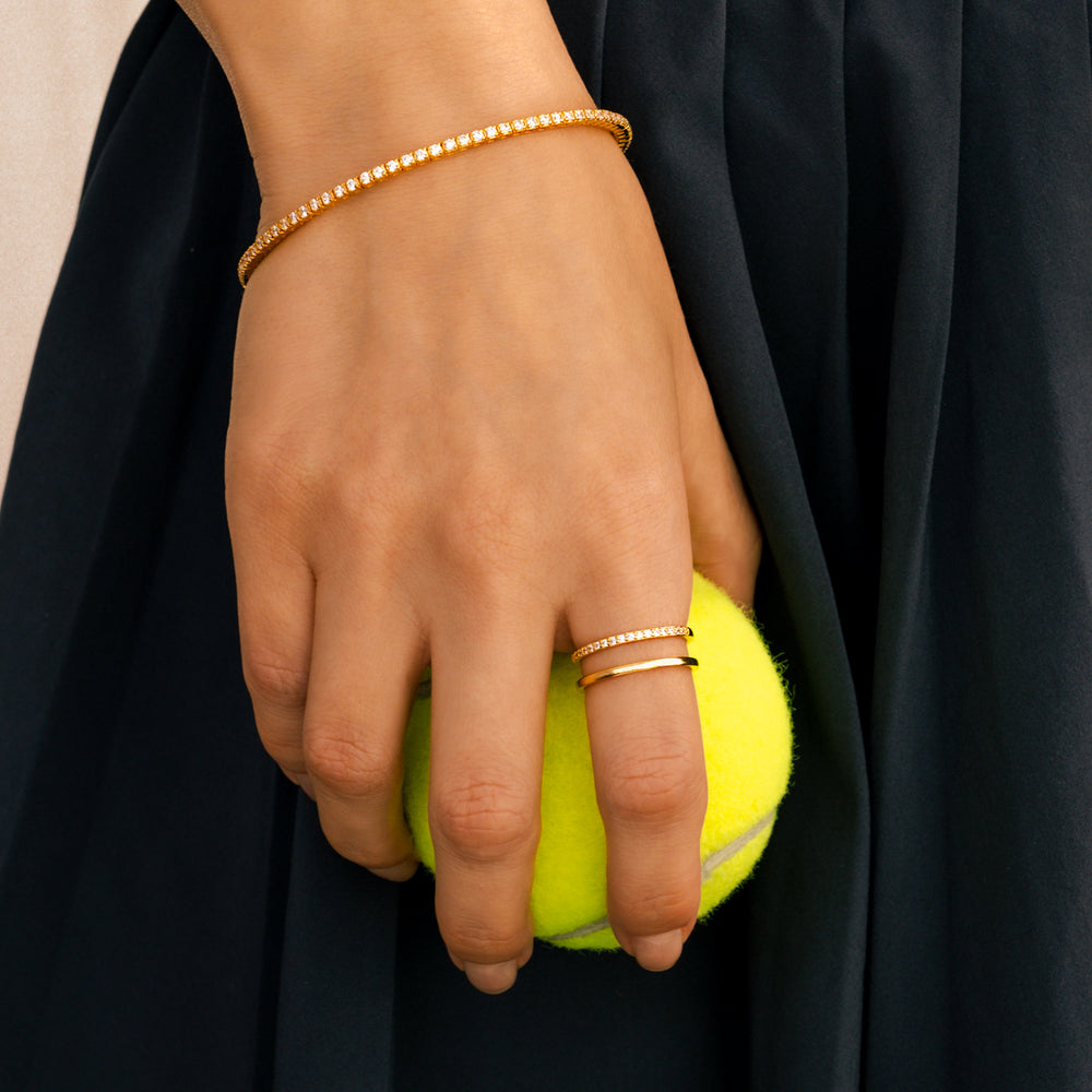 Hand holding a yellow tennis ball with gold bracelets and rings against a black background