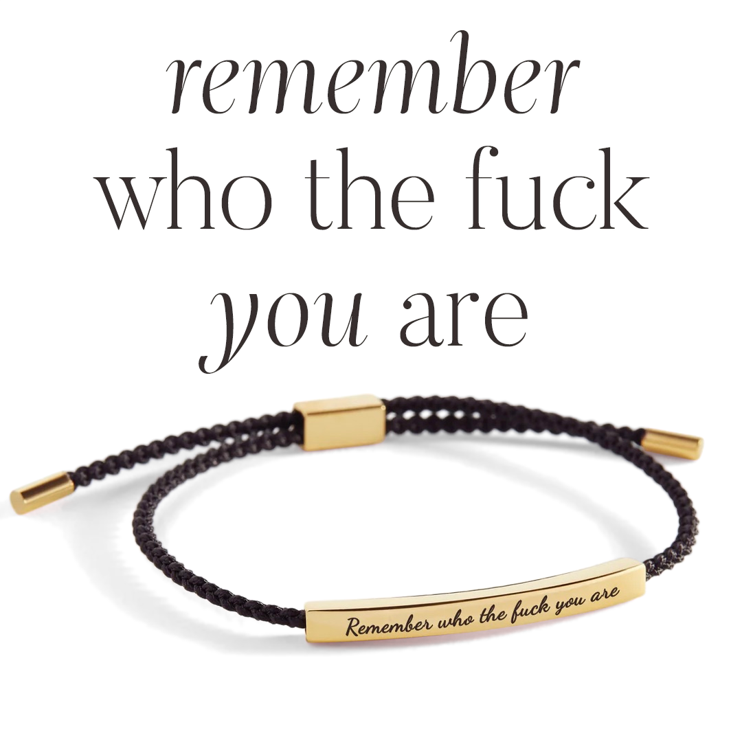 A braided black nylon bracelet with gold-plated ends and a gold-plated bar charm with the inscription 'remember who the f*** you are'. The image has a white background.