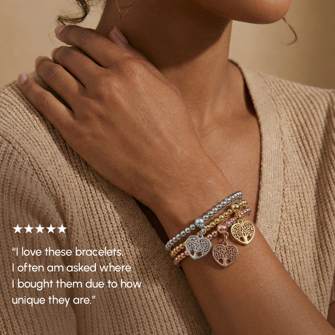 Woman's arm wearing multiple bracelets with heart and tree charms on a beige background